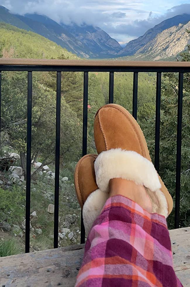 Reviewer wearing the chesnut colored slippers successful beforehand of a upland backdrop