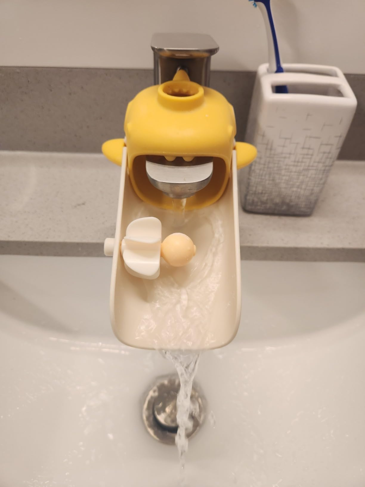 Yellow sink faucet extender with fun design, promoting easy hand washing for kids. Water flows down the spout into a bathroom sink