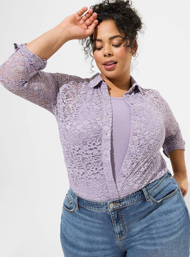A person poses in a purple lace shirt and denim jeans, looking to the side