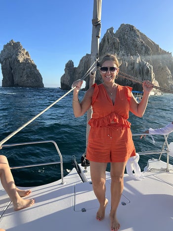 Woman in a casual orange romper smiles on a boat with rocky landscape in background