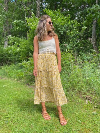 Person in a sleeveless top, floral skirt, and sandals stands on grass with a leafy background, wearing sunglasses and smiling