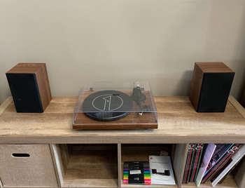 Reviewer image of two wooden and black bookshelf speakers next to matching turntable with clear cover and black needle and dials on top of wooden bookshelf