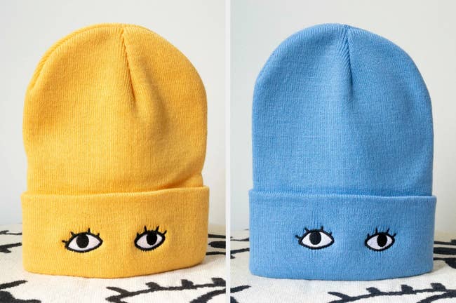 Two knit beanies with embroidered black and white eyes in yellow and light blue standing up on an eye patterned carpet