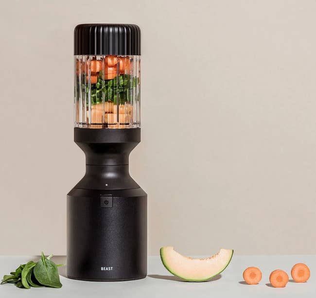 the black blender full of fruits and vegetables with spinach, melon, and carrots on the ground next to it