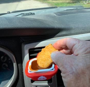 The dip clip clipped onto a reviewer's car air vent with sauce and reviewer dipping nugget in it