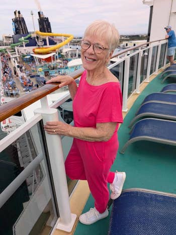 Person in a stylish jumpsuit and sneakers smiles on a cruise ship deck, with water slides and tourists in the background