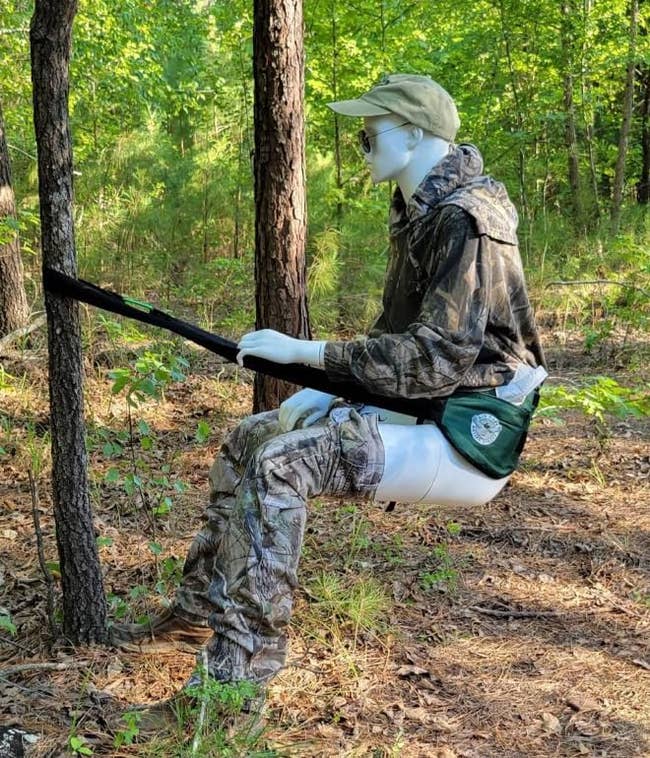 Mannequin dressed in camouflage gear using a strep mounted to a tree to sit in a squatted position, suitable for outdoor bathroom use 