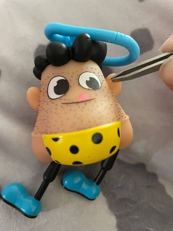 Toy keychain resembling a caveman with cartoonish features, being held by tweezers