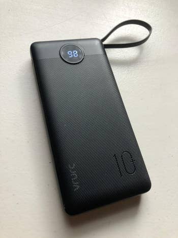 Portable charger with digital display showing 98% charge, a textured surface, and a loop for carrying