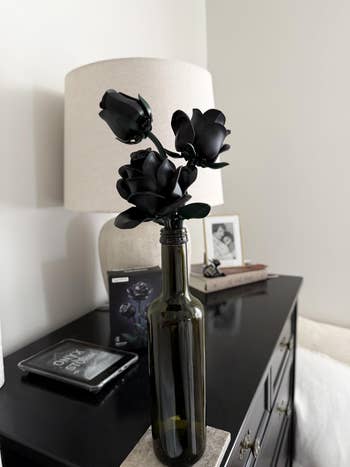 Black roses successful a vino vessel vase connected a dresser with a lamp, framed photo, and perfume container nearby. Modern location decor style