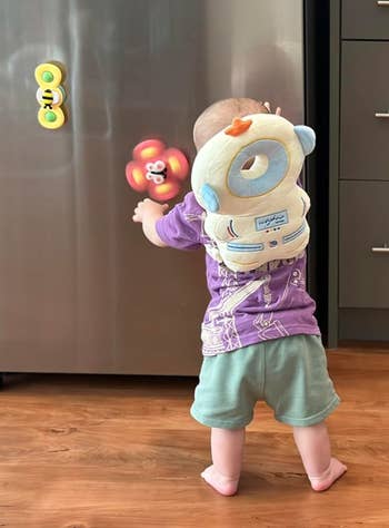 A baby wearing a plush astronaut backpack stands in front of a refrigerator, reaching for colorful magnetic toys attached to the fridge door