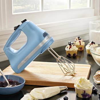 A airy  bluish  manus  mixer connected  a room  antagonistic  with cupcakes, icing bags, and berries nearby, suggesting a baking scene