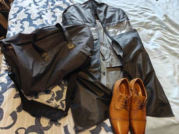 A neatly arranged display of a gray suit inside a black garment bag, brown dress shoes, and a gray duffel bag on a patterned bedspread