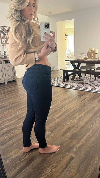 A reviewer taking a mirror selfie showcasing their casual outfit of a beige sweater and dark jeans, with bare feet, in a cozy, modern home setting