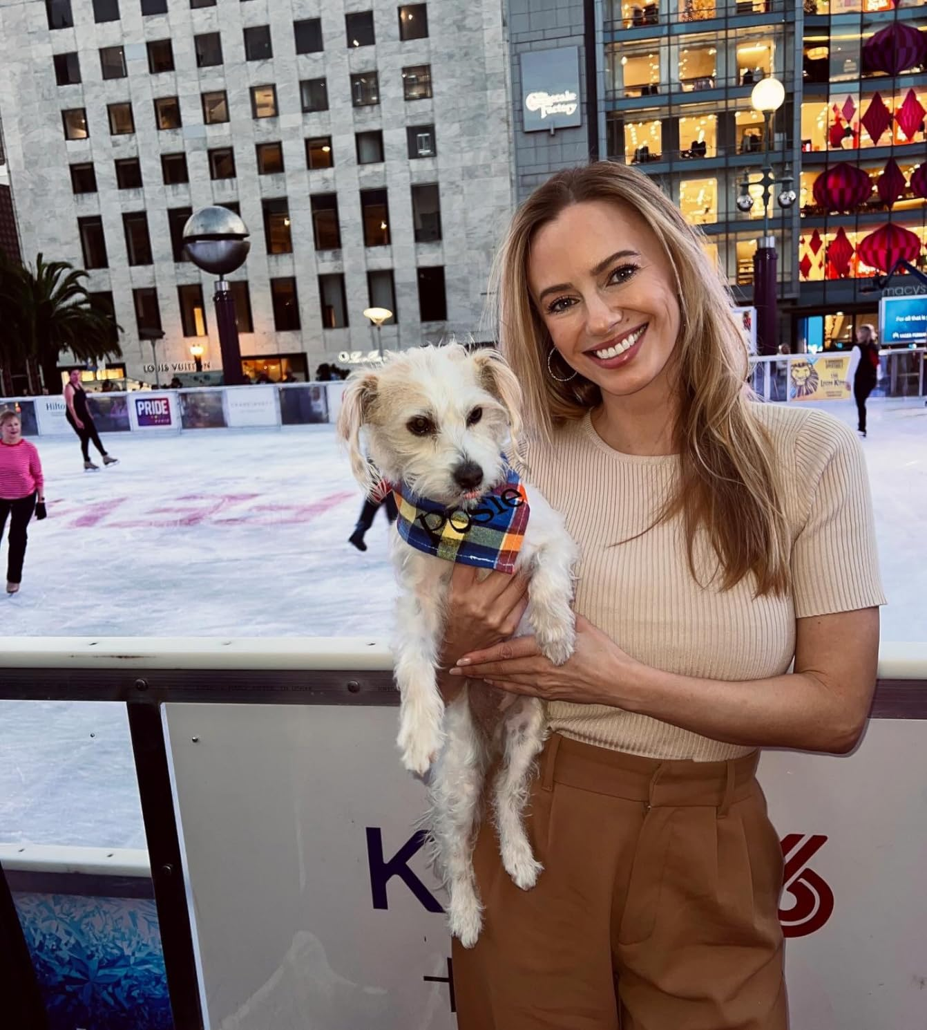Person holding a small dog in front of an outdoor skating rink, wearing a casual beige top and brown pants. Urban backdrop with shops