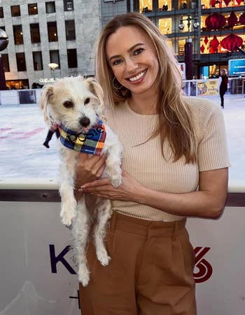 Person holding a small dog in front of an outdoor skating rink, wearing a casual beige top and brown pants. Urban backdrop with shops