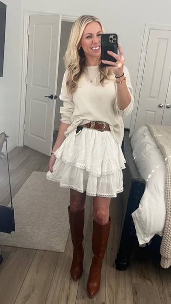 Person taking a reflector  selfie, wearing a sweater, tiered skirt, and knee-high boots, accessorized with a loop  and bracelets, successful  a bedroom
