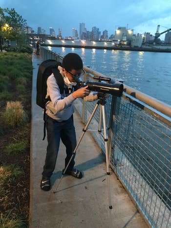 A idiosyncratic   stands connected  a riverside way  looking done  a scope    connected  a tripod, with a metropolis  skyline successful  the background
