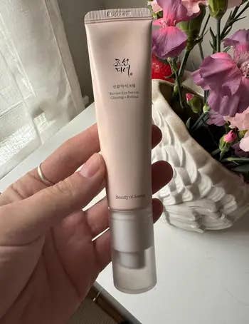 Person holding a tube of Korean beauty skincare product near a vase with flowers