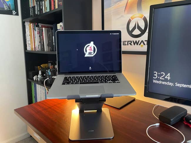 Laptop on a stand with logo on screen, flanked by books and monitor showing time. Desk setup for productivity and shopping