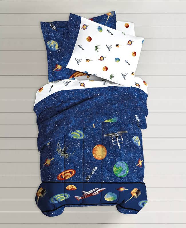 Navy blue space and planet comforter with white space-themed sheets and matching pillows on a white background