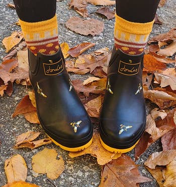 Person in black boots with bee design, standing on autumn leaves. Featured in a shopping category