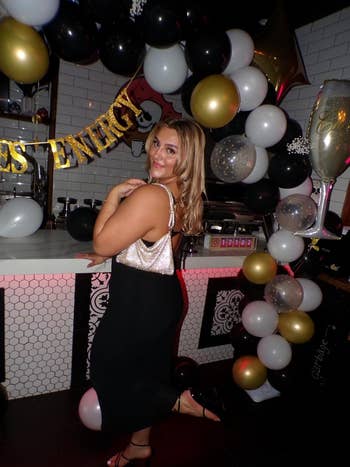 Person in a shiny top and black skirt poses at a festive event with balloon decor and a champagne glass prop, suggesting a celebratory occasion
