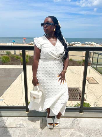 Person in a ruffled white dress with textured patterns, wearing sunglasses, stands on a beachside terrace with a small white bag and platform shoes