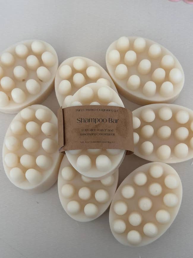 Oval shampoo bars with textured dots are stacked. One has a brown paper label indicating it contains shea butter and essential oils