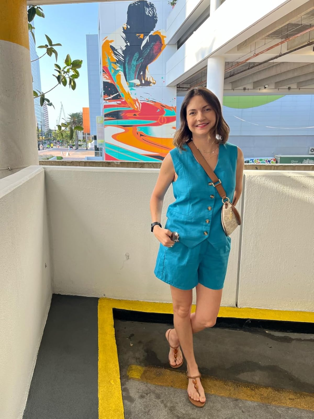 Person successful  a sleeveless, button-up bluish  jumpsuit and sandals stands successful  a parking lot, with a colorful mural successful  the background