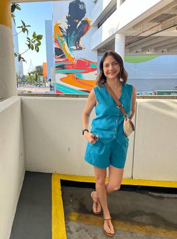 Person successful a sleeveless, button-up bluish jumpsuit and sandals stands successful a parking lot, with a colorful mural successful the background