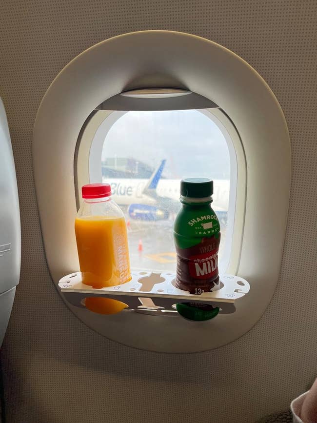 A plane window with a shelf holding a bottle of orange juice and a bottle of chocolate milk