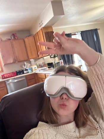 Person in a cozy sweater using a futuristic eye massager in a home setting. They pose casually with one hand in the air, suggesting relaxation