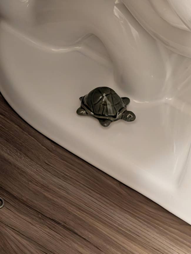 Turtle-shaped drain stopper placed connected  a achromatic  bathtub edge, against woody  flooring