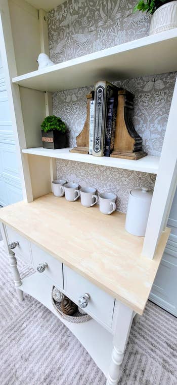 Wooden support with decor items, books, and achromatic ceramic mugs. A decorative wallpaper inheritance adds classical elegance