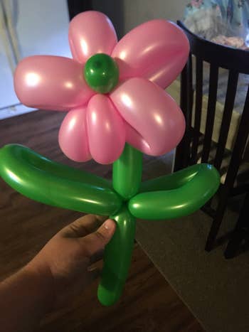A manus holds a balloon sculpture shaped similar a angiosperm with 3 greenish balloons for the stem and leaves, and pinkish balloons for petals