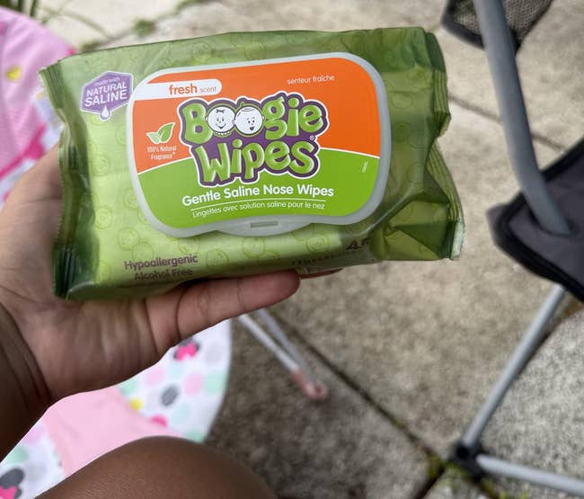 Person holding a battalion of Boogie Wipes, gentle saline chemoreceptor wipes, outdoors with chairs and factual patio visible