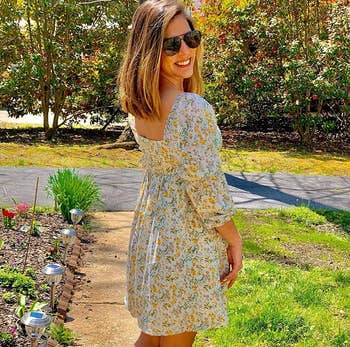 Woman in casual floral dress poses outdoors, smiling, with one hand on hip, promoting spring fashion