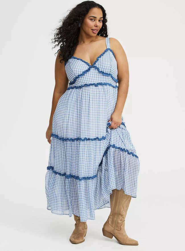 A person in a long checkered dress with lace trim and cowboy boots poses confidently, showcasing a stylish and casual look suitable for shopping