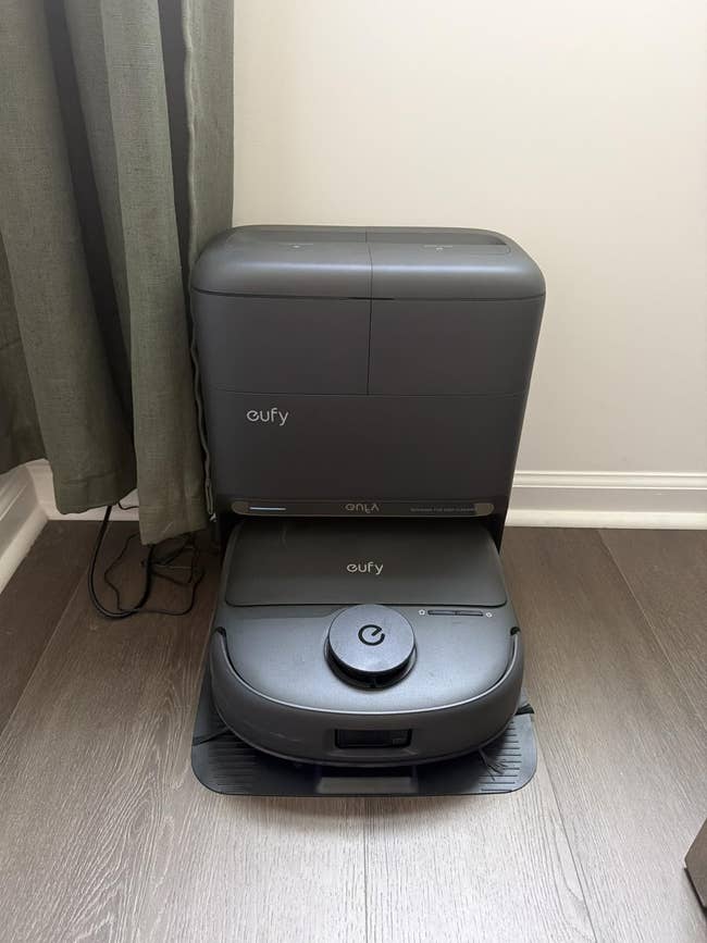 A robotic vacuum cleaner docked connected  its charging station, placed connected  a hardwood level  adjacent  to a curtain