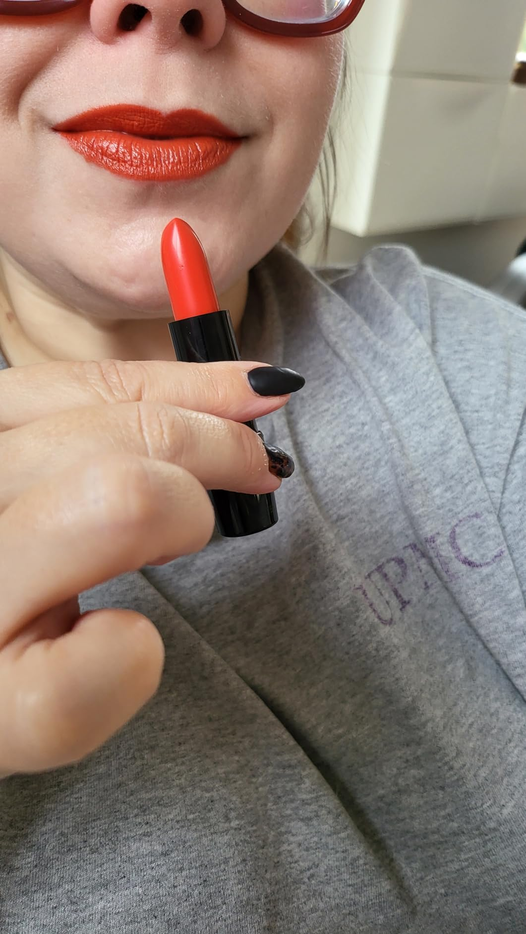 Person wearing sunglasses applies reddish  lipstick, holding it adjacent   their lips. They deterioration  a casual top