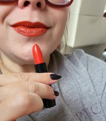 Person wearing sunglasses applies reddish lipstick, holding it adjacent their lips. They deterioration a casual top