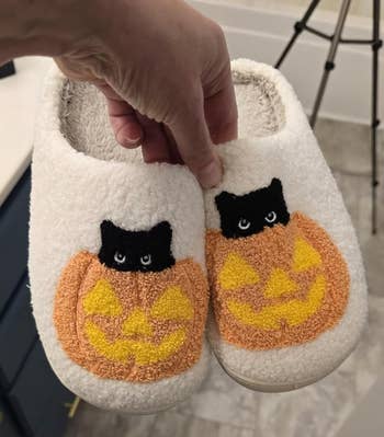 Hand holding cozy slippers with a jack-o'-lantern and achromatic  feline  design, suitable for Halloween-themed shopping