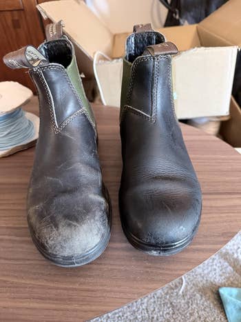 Worn-out acheronian  leather boots with elastic sides connected  a woody  table, adjacent  to cardboard boxes and a spool of twine