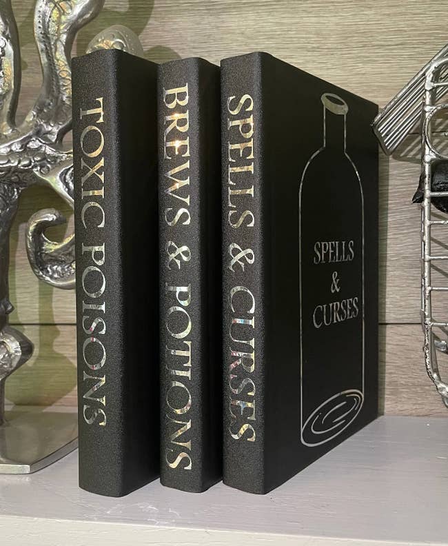 Three black books with holographic words on the spines that say: Toxic Poisons, Brews & Potions, Spells & Curses