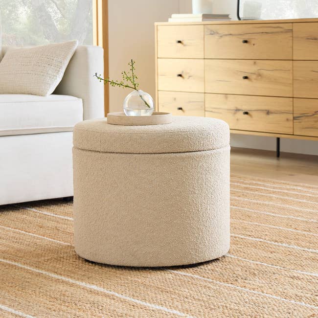 The storage ottoman in a light colored tweed fabric with a small plant in a vase on top