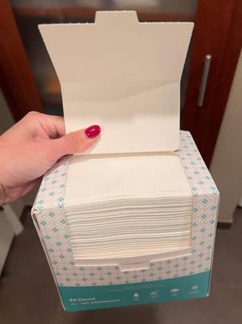 Hand holding an unfastened container of look cleansing wipes successful a bathroom, showing a stack of wipes wrong the carton