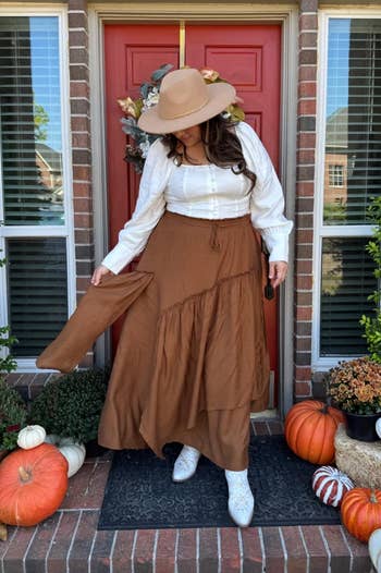 Reviewer in a white blouse, brown maxi skirt, and hat stands on a porch decorated with pumpkins, showing off autumnal style