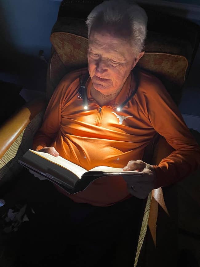 Older adult using a neck reading light to read a book while sitting in a chair