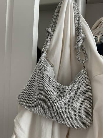 Silver, rhinestone-studded handbag with knotted straps, hanging on a white surface, showcasing a chic, glamorous style suitable for shopping